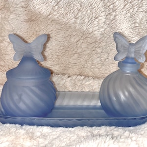 Peut inclure: Un ensemble de deux flacons en verre givré avec des bouchons en forme de papillon, sur un plateau assorti. Les flacons sont de couleur bleu clair et présentent un motif en spirale.