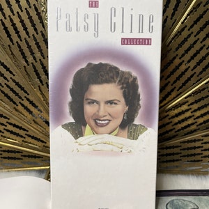 Puede incluir: Una caja de CD blanca con una foto de Patsy Cline en la parte delantera. El texto "The Patsy Cline Collection" está impreso en plata en la caja.
