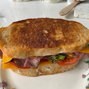 May include: A toasted sandwich on a floral plate. The sandwich features layers of roast beef, cheddar cheese, lettuce, and tomato between two slices of golden-brown bread. The bread has a crispy, textured crust.