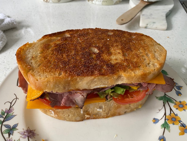 May include: A toasted sandwich on a floral plate. The sandwich features layers of roast beef, cheddar cheese, lettuce, and tomato between two slices of golden-brown bread. The bread has a crispy, textured crust.