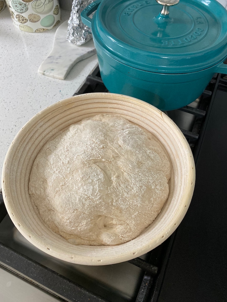 May include: A round, light-colored banneton basket holds a ball of dough dusted with flour, ready for baking. A teal-colored Dutch oven sits nearby. The scene suggests homemade bread baking.