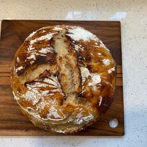 May include: A round loaf of freshly baked bread with a golden-brown crust, dusted with flour, sits on a wooden cutting board. The bread has a rustic appearance with visible scoring and a slightly uneven surface.