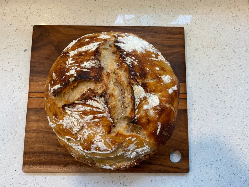 May include: A round loaf of freshly baked bread with a golden-brown crust, dusted with flour, sits on a wooden cutting board. The bread has a rustic appearance with visible scoring and a slightly uneven surface.