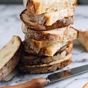 May include: A stack of freshly sliced bread with a crusty exterior and soft interior. The bread slices are stacked on top of each other, with a knife with a wooden handle resting on the surface next to the bread. The bread is light brown and beige.