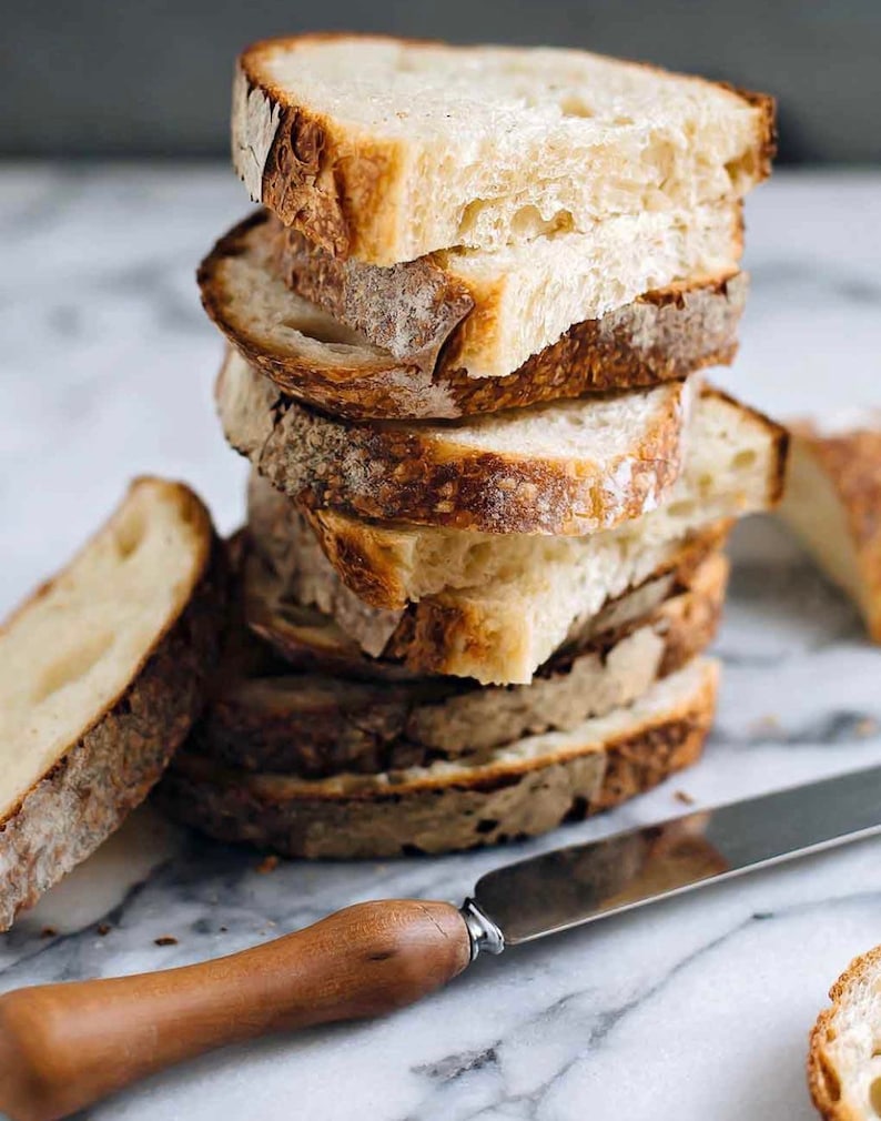 May include: A stack of freshly sliced bread with a crusty exterior and soft interior. The bread slices are stacked on top of each other, with a knife with a wooden handle resting on the surface next to the bread. The bread is light brown and beige.