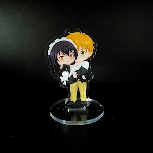 May include: A clear acrylic stand with a cartoon illustration of a couple. The male character has blonde hair and is holding a female character with black hair. The female character is wearing a maid outfit.
