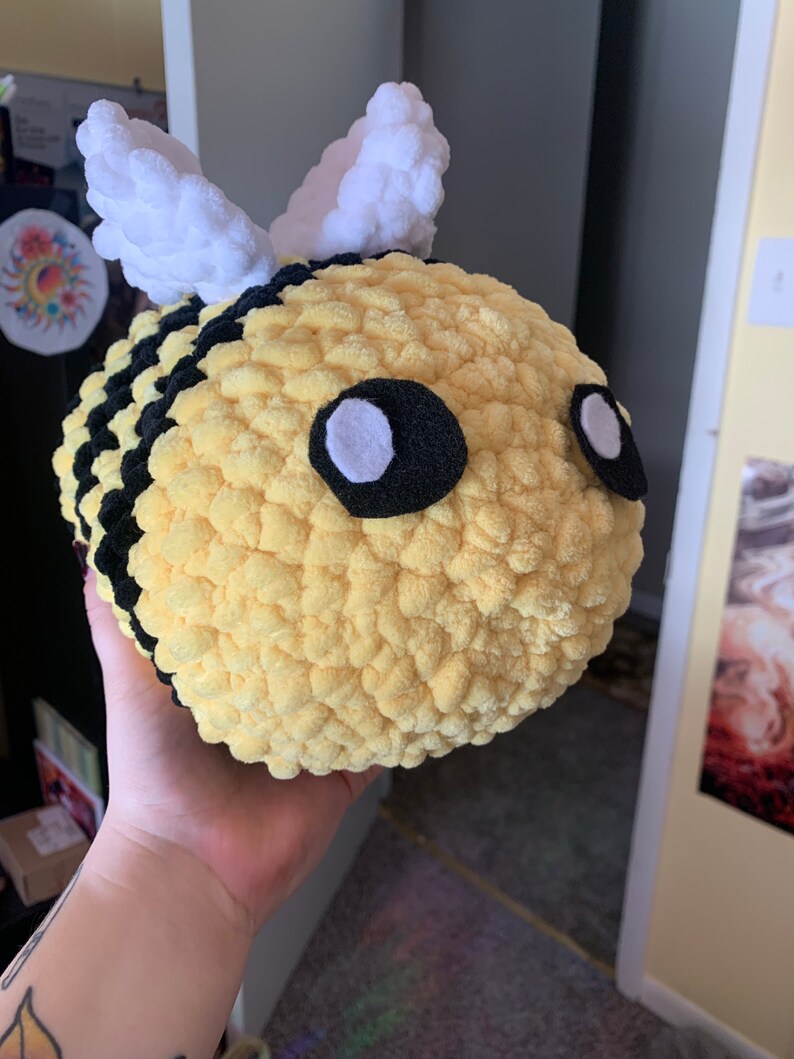 Pode incluir: Uma abelha de pel&uacute;cia feita &agrave; m&atilde;o com corpo amarelo, listras pretas e asas brancas. A abelha tem olhos grandes e redondos com feltro preto e centros brancos. A pel&uacute;cia &eacute; macia e texturizada, perfeita para o quarto de uma crian&ccedil;a ou como item decorativo.