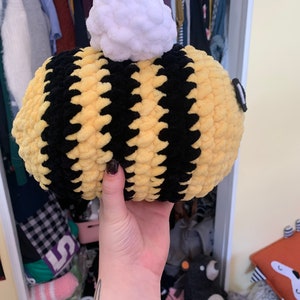 Pode incluir: Uma abelha de pel&uacute;cia feita &agrave; m&atilde;o, com listras amarelas e pretas. A abelha tem um pompom branco no topo e um pequeno olho preto. A abelha &eacute; segurada em uma m&atilde;o.