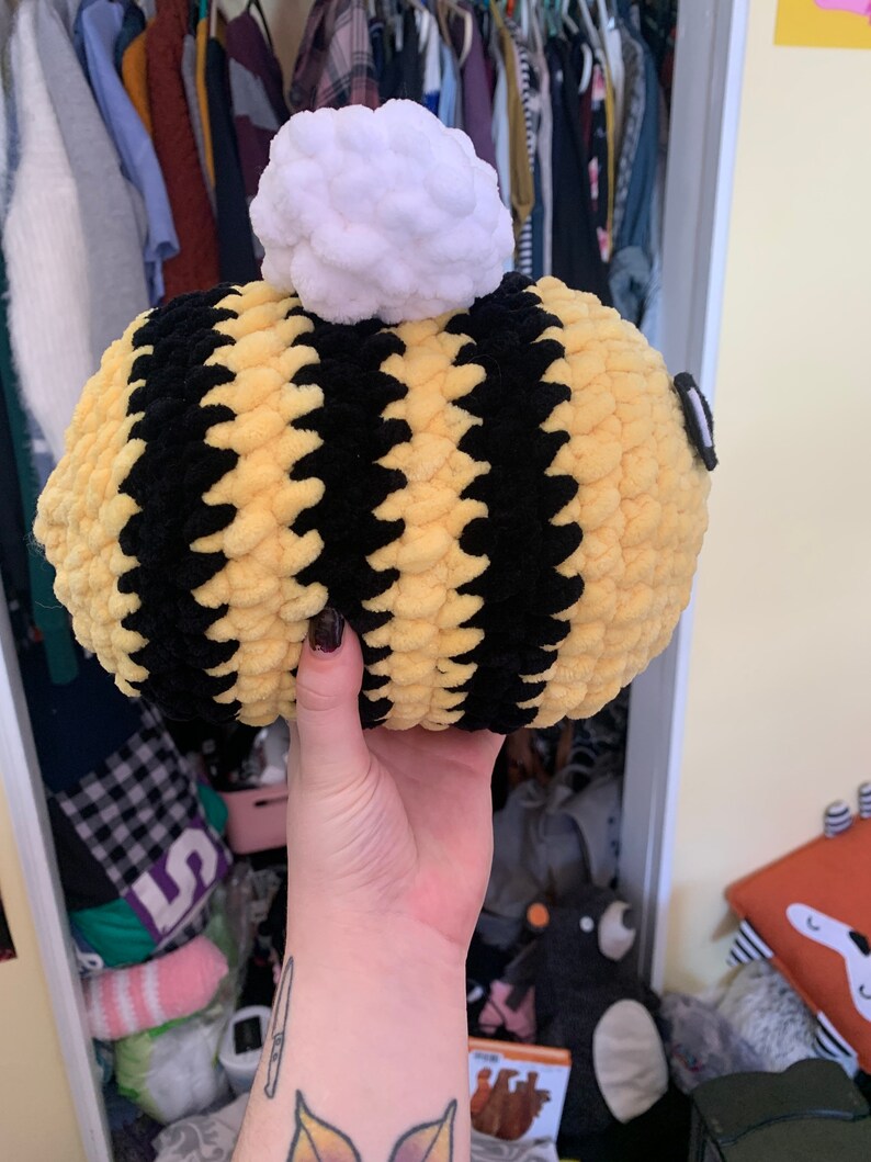 Pode incluir: Uma abelha de pel&uacute;cia feita &agrave; m&atilde;o, com listras amarelas e pretas. A abelha tem um pompom branco no topo e um pequeno olho preto. A abelha &eacute; segurada em uma m&atilde;o.