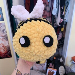 May include: A handmade, plush bee toy with a yellow body, black stripes, and white wings. The bee has large, round eyes with black outlines and white centers. The toy is soft and round, perfect for a child's room or as a decorative item.