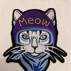 May include: Embroidered patch of a white cat wearing a purple helmet with the word "Meow" on it and blue goggles. The cat is wearing a blue bandana.