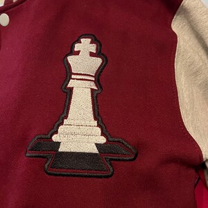 Puede incluir: Una chaqueta de béisbol de color granate y gris con un rey de ajedrez bordado en blanco en el lado izquierdo de la chaqueta.