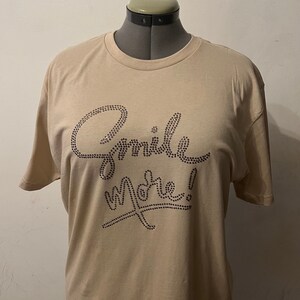 May include: A beige t-shirt with the words "Smile More!" in black rhinestones.
