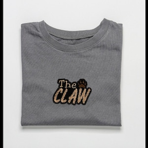 May include: A heather grey t-shirt with a black and brown embroidered patch that says "The CLAW" with a paw print.