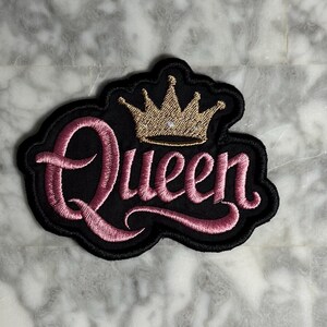 May include: Black patch with the word "Queen" in pink script, outlined in black. A gold crown with rhinestones sits above the word. The patch has a black border.