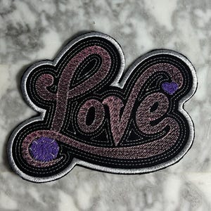 May include: Embroidered patch with the word "Love" in a cursive font. The patch features a black background with a pink and purple gradient, outlined in white and black stitching. A small purple heart and circle accent the design.