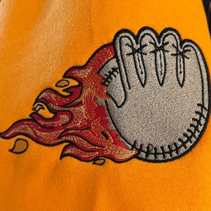 以下が含まれることがあります： 炎に包まれた刺繍された野球グローブがついた黄色のスウェットシャツ。グローブはグレーで、炎は赤とオレンジです。