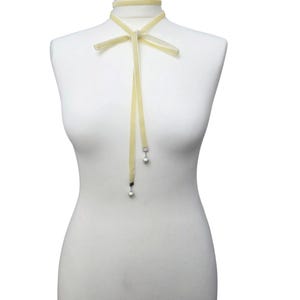 Pu&ograve; includere: Una collana girocollo in velluto giallo pallido con fiocco e nastri pendenti. I nastri terminano con piccole perle argentate pendenti. La collana &egrave; esposta su un manichino.