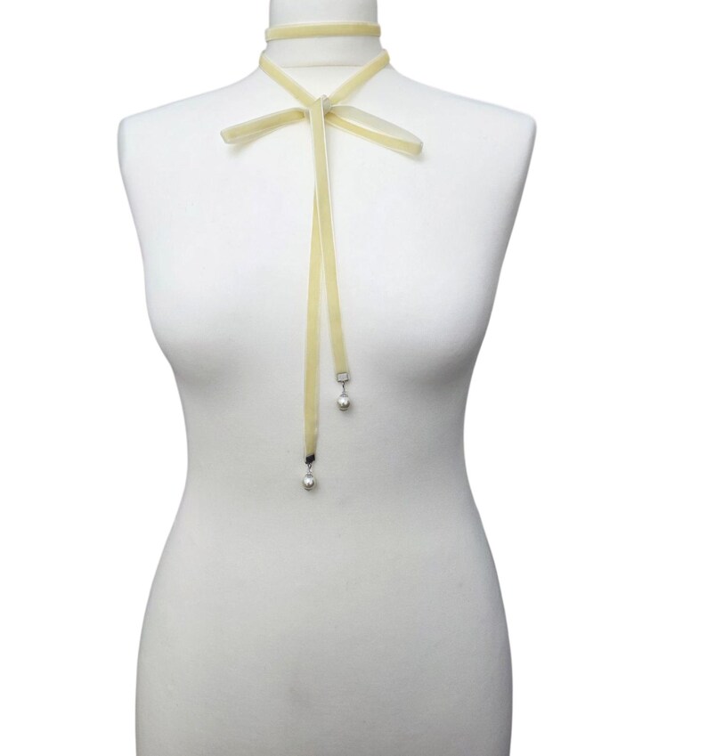 Pu&ograve; includere: Una collana girocollo in velluto giallo pallido con fiocco e nastri pendenti. I nastri terminano con piccole perle argentate pendenti. La collana &egrave; esposta su un manichino.