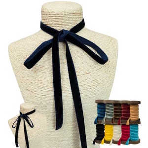 May include: A dark navy blue velvet ribbon tied in a simple knot around a beige mannequin neck. The ribbon features a soft, plush texture and long, dangling ends. A collection of various coloured velvet ribbons on wooden spools is also visible.