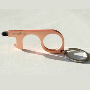 May include: Rose gold-colored keychain tool with a stylus, bottle opener, and two loops for touching surfaces. The keychain is engraved with the name "Christina".