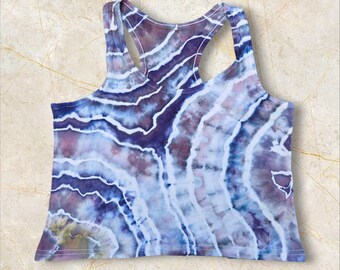 Pode incluir: Uma regata tie-dye com costas de nadador em tons de azul, roxo e branco. O padrão apresenta desenhos ondulados e listrados. A regata tem decote redondo e design sem mangas.