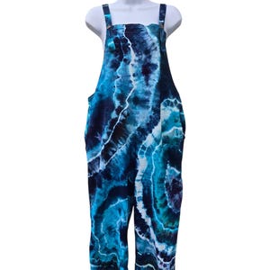 Könnte beinhalten: Blau gefärbte Latzhose mit verstellbaren Trägern und Holzknöpfen. Das Kleidungsstück hat ein lebendiges Muster aus Blau und Weiß, das einen fließenden Aquarelleffekt erzeugt. Die Latzhose wird auf einer weißen Schaufensterpuppe präsentiert.