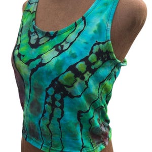 Pode incluir: Um top sem mangas tie-dye em tons de verde-azulado, verde e preto. O top sem mangas apresenta um decote redondo e um comprimento curto. O tecido tem um efeito marmorizado com padrões escuros de forma orgânica. A peça de roupa é exibida num manequim.