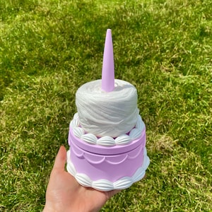 May include: A two-tiered cake-shaped yarn holder with a white yarn ball on top and a purple base. The holder has white decorative details around the edges.