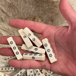 May include: A collection of white bone dice with black circles on each side. The dice are in a hand, and there are more dice in the background.