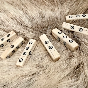 May include: Seven bone dice with black circles on each side. The dice are arranged in a semi-circle on a light brown fur surface.