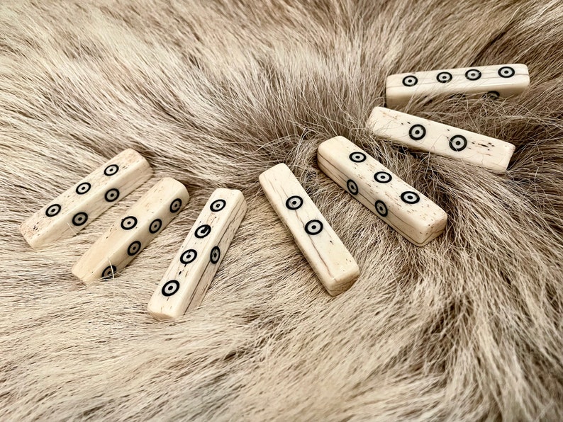 May include: Seven bone dice with black circles on each side. The dice are arranged in a semi-circle on a light brown fur surface.