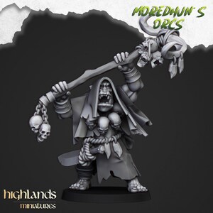 Könnte beinhalten: Eine graue Plastikminiatur eines Orc-Schamanen, der eine Kapuzenrobe trägt und einen Stab mit Schädeln hält. Der Orc hat einen großen Mund und einen Ring in der Nase. Der Stab hat eine Kette mit Schädeln, die daran hängen. Die Miniatur ist Teil der "Moredhun's Orcs"-Kollektion.