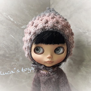 Könnte beinhalten: Eine Puppe trägt eine rosa und graue Häkelmütze mit einem strukturierten Design. Die Mütze hat eine spitze Spitze und bedeckt die Ohren der Puppe. Die Puppe hat schwarze Haare und große, blaue Augen. Die Puppe trägt einen grauen Pullover.