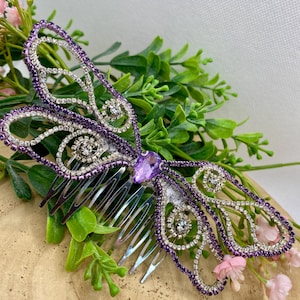 May include: A hair comb featuring a butterfly-shaped ornament decorated with purple and clear rhinestones. The center of the butterfly is accented with a single, teardrop-shaped purple crystal, and the wings are outlined in purple and clear stones.