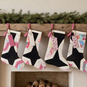 May include: Four quilted Christmas stockings hanging in a row. Each stocking features a large black star shape centered on a patchwork background of various floral and checkered fabrics in shades of red, blue, and white. Each has a small fabric tag.