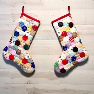 May include: Two Christmas stockings made from a patchwork of multicolored fabric hexagons. Each stocking features a bright red fabric cuff at the top with a matching red hanging loop. The quilted surface displays a variety of patterns and colors.