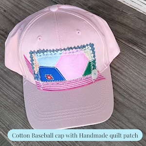 May include: A light pink baseball cap featuring a rectangular handmade quilt patch on the front. The patch consists of multi-coloured fabric segments in blue, pink, and green, accented with a small white button and a pink and white gingham trim.