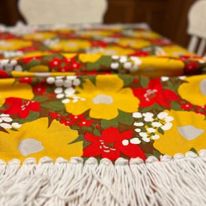 May include: A rectangular fabric tablecloth featuring a vibrant floral pattern with large yellow and red flowers, small white blossoms, and green foliage on a brown background, finished with a thick white fringe trim along the bottom edge.