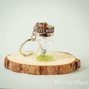 Peut inclure: Un petit porte-clés en forme de bouteille en verre avec un bouchon en liège sur lequel est inscrit "Hey Listen!"  À l'intérieur de la bouteille se trouve une petite fée bleue avec de l'herbe verte au fond.