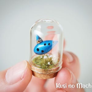 May include: A small glass jar with a cork lid contains a blue ocarina with a silver cap, surrounded by green moss and white flowers.