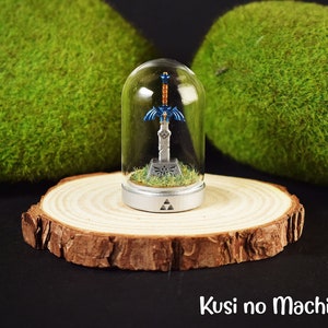 May include: A miniature Master Sword from The Legend of Zelda video game series is displayed in a glass dome on a wooden base. The sword is on a pedestal with a Triforce symbol. The base is surrounded by green moss.