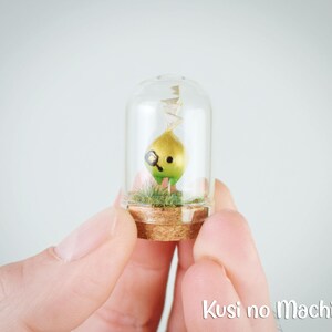 Puede incluir: Una pequeña figurita verde y dorada con un solo ojo y una sonrisa está dentro de un frasco de vidrio con un tapón de corcho. La figurita está sentada sobre un lecho de musgo verde. El frasco está siendo sostenido por una mano. El texto "Kusi no Machi" es visible en la parte inferior de la imagen.