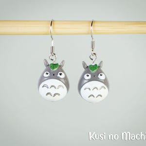 Könnte beinhalten: Ein Paar grau-weiße Totoro-Ohrringe mit silbernen Haken. Jeder Ohrring hat ein grünes Blattdetail. Die Ohrringe werden vor einem hellgrauen Hintergrund präsentiert.