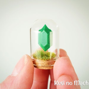 Könnte beinhalten: Ein kleines Glasgefäß mit einem Korkdeckel enthält ein grünes, geometrisch geformtes Objekt und ein kleines Stück grünes Moos. Der Text "Kusi no Machi" ist unter dem Gefäß gedruckt.
