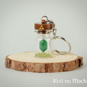 May include: A green gemstone charm inside a glass bottle with a brown lid featuring the Triforce symbol. The bottle is attached to a silver key chain and is sitting on a wooden surface. Kusi no Machi is written below the bottle.