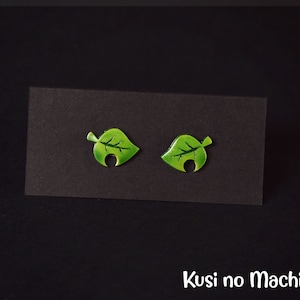 May include: A pair of green leaf-shaped earrings with a hole in the centre of each leaf. The earrings are on a black background.