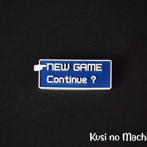 May include: A blue and white pixelated button with the text "NEW GAME Continue?" on it. The button is a 3D printed replica of a video game menu.