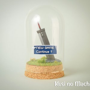 Op de afbeelding: Een miniatuurreplica van het Buster Sword uit Final Fantasy VII, ingesloten in een glazen koepel op een kurken basis met een klein stukje groen gras. Het zwaard is gedeeltelijk bedekt door een blauw-wit bord met de tekst "NEW GAME Continue?"