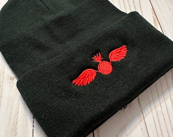 Puede incluir: Gorro negro con un diseño bordado en rojo de una esfera alada. El gorro está hecho de un material suave y tejido y tiene un borde doblado. El diseño bordado está centrado en el borde doblado.